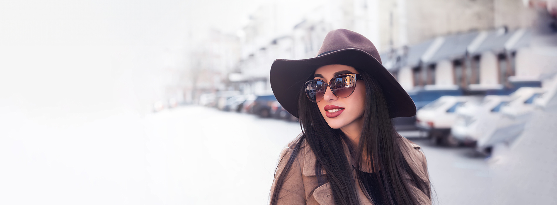 A woman in a hat and sunglasses is standing on a street, with a blurred background that includes buildings and cars.