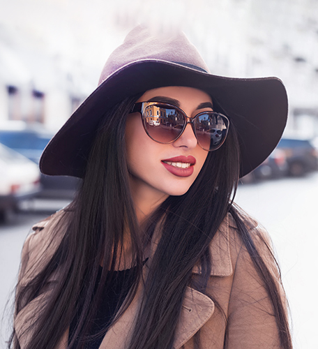 A woman with long dark hair, wearing sunglasses and a brown hat, stands on a city street in a coat and heels.