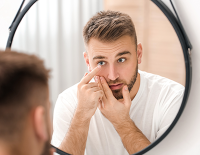 A man in a white t-shirt is seated in front of a round mirror, looking at his reflection while applying a product to his skin with both hands.