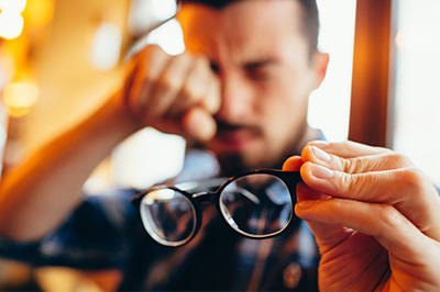 The image shows a man holding a pair of glasses, with his hand on the frame as he looks at them.