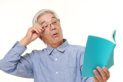 An elderly man with glasses, holding a book and looking intently at it.