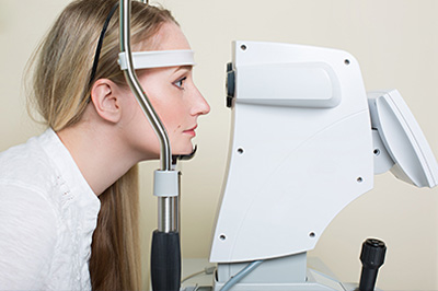 A woman with blonde hair is wearing a headset and looking through an ophthalmoscope machine, which is placed on a stand.