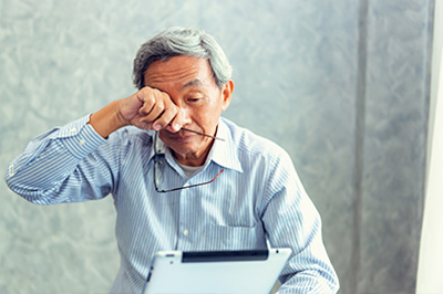 An elderly Asian man is sitting at a desk with his head tilted back, holding a tissue to his nose as he looks upwards while using a tablet computer.