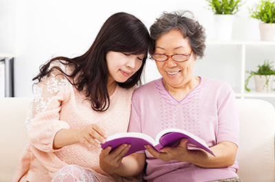 An elderly person and a younger individual are sitting together, both engrossed in reading a book.