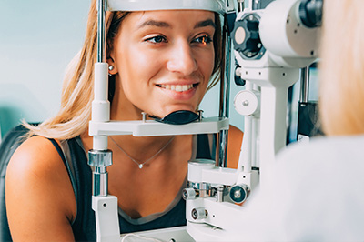 An adult woman in a medical setting, smiling at the camera while seated in an eye exam chair with a device attached to her face.