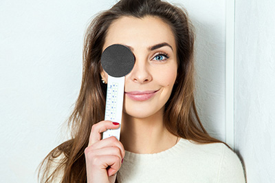 A woman holds a thermometer up to her forehead, with a magnifying glass placed over her eye.